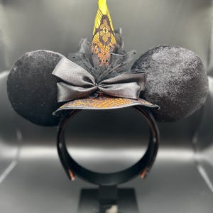 May include: Black mouse ear headband with a witch hat design. The ears are round and covered in black velvet. The witch hat is orange and black with a lace overlay, a black bow, and a yellow tip. The headband is black.