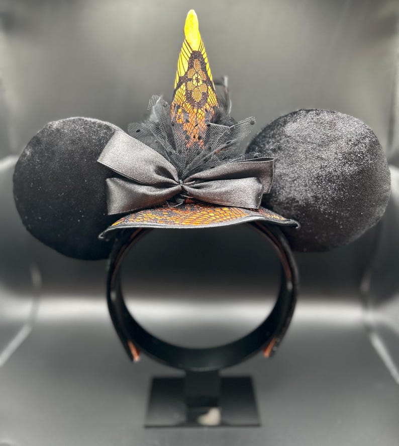 May include: Black mouse ear headband with a witch hat design. The ears are round and covered in black velvet. The witch hat is orange and black with a lace overlay, a black bow, and a yellow tip. The headband is black.