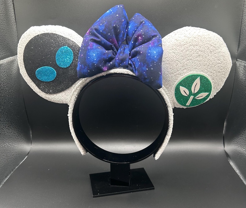 May include: A whimsical headband featuring two white sequined ears, one with a black face and blue glitter accents, the other with a green leaf emblem. A large blue galaxy-print bow sits atop the headband.