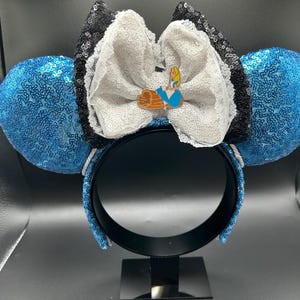 May include: A whimsical headband featuring blue sequined ears. A large white bow with black sequin trim and a small Alice in Wonderland pin adorns the center. The headband is black.