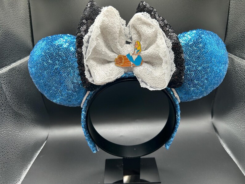 May include: A whimsical headband featuring blue sequined ears. A large white bow with black sequin trim and a small Alice in Wonderland pin adorns the center. The headband is black.