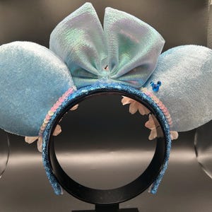 Peut inclure: Un serre-t&ecirc;te avec deux grandes oreilles en velours bleu clair et un n&oelig;ud central bleu clair iris&eacute;. L'arceau est recouvert de sequins bleu fonc&eacute;, agr&eacute;ment&eacute; d'une rang&eacute;e de sequins rose p&acirc;le et de petites fleurs blanches &agrave; la base.