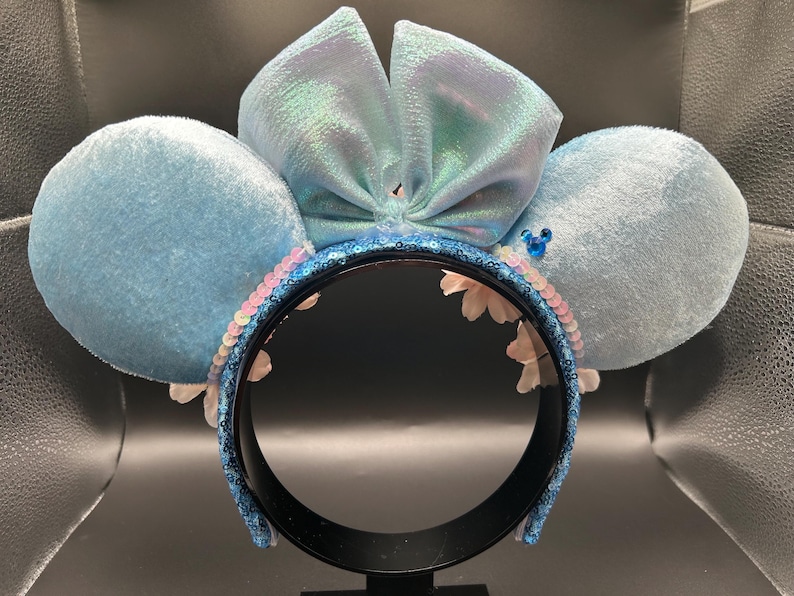 Peut inclure: Un serre-t&ecirc;te avec deux grandes oreilles en velours bleu clair et un n&oelig;ud central bleu clair iris&eacute;. L'arceau est recouvert de sequins bleu fonc&eacute;, agr&eacute;ment&eacute; d'une rang&eacute;e de sequins rose p&acirc;le et de petites fleurs blanches &agrave; la base.