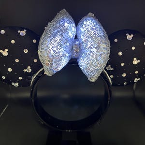 May include: Black mouse ear headband with a silver sequined bow and black ears. The ears are decorated with clear rhinestones and small Mickey Mouse silhouettes. The bow is illuminated with white lights.