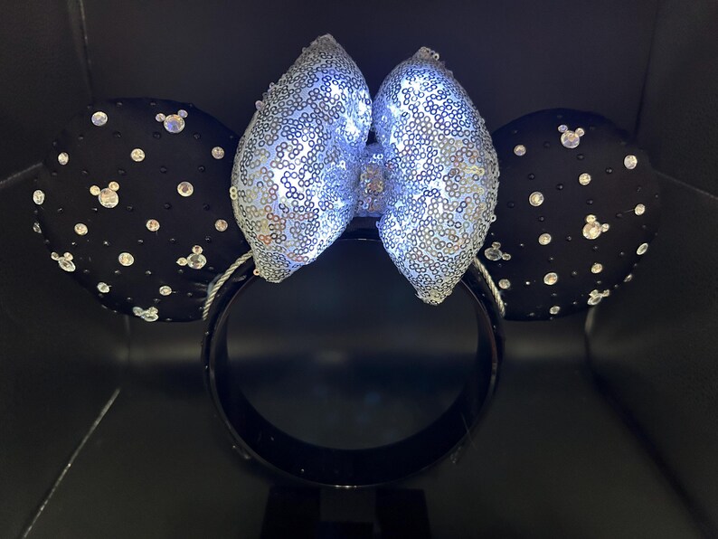 May include: Black mouse ear headband with a silver sequined bow and black ears. The ears are decorated with clear rhinestones and small Mickey Mouse silhouettes. The bow is illuminated with white lights.