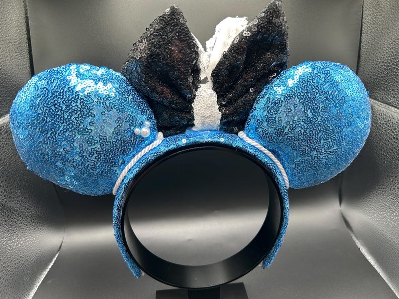 May include: A pair of mouse ears with blue sequined ears and a black sequined bow with white accents. The headband is black with a white rope detail. A fun accessory for themed events.