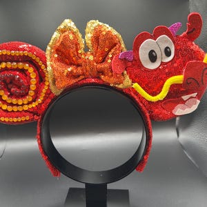 May include: A red sequined headband with a dragon design. The headband features a bow with orange sequins and a dragon head with white eyes, a yellow mustache, and a red mouth. The ears are red with a spiral design.