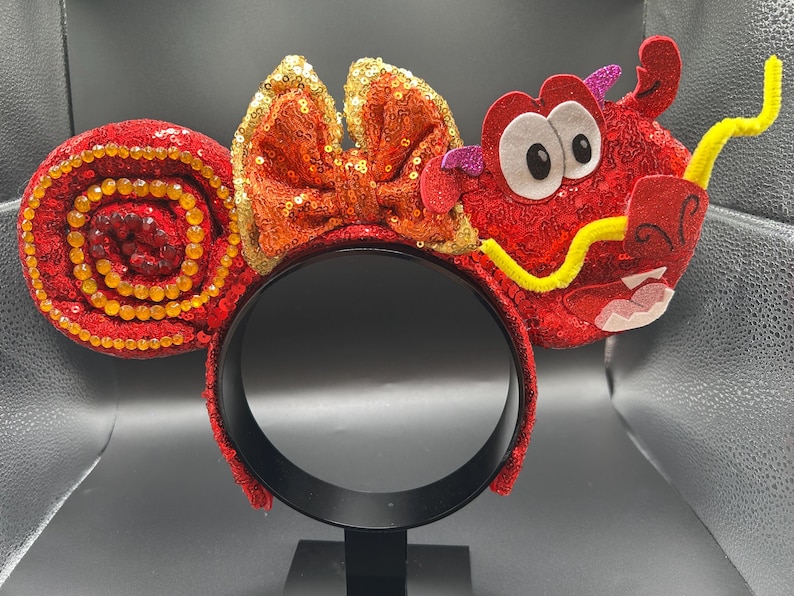 May include: A red sequined headband with a dragon design. The headband features a bow with orange sequins and a dragon head with white eyes, a yellow mustache, and a red mouth. The ears are red with a spiral design.