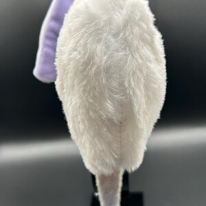 May include: A plush, white, furry headband with a lavender accent. The headband is designed with a soft, textured material, and is likely for a costume or themed event. The headband is displayed on a black stand against a dark background.