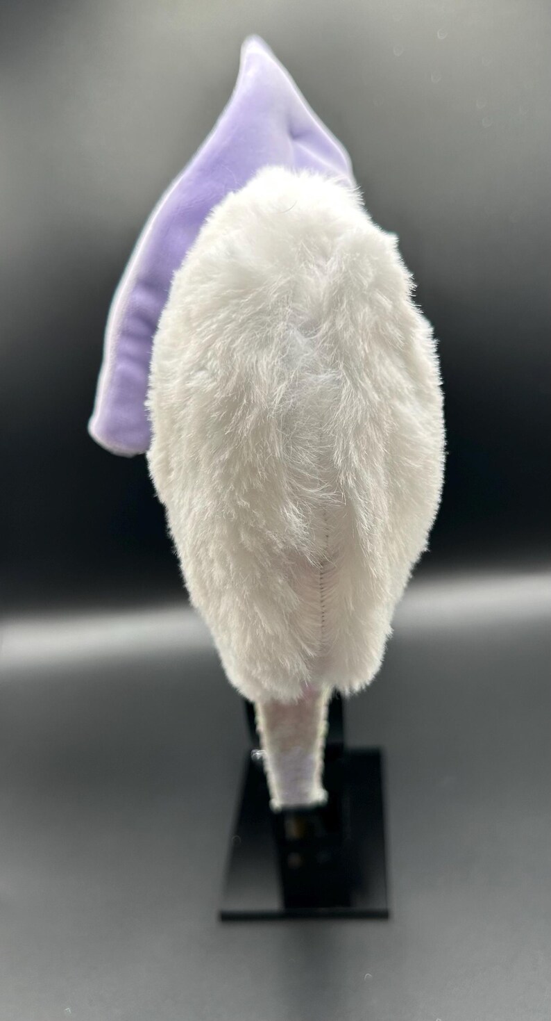 May include: A plush, white, furry headband with a lavender accent. The headband is designed with a soft, textured material, and is likely for a costume or themed event. The headband is displayed on a black stand against a dark background.