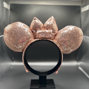 May include: Rose gold sequined mouse ear headband with a matching bow. The ears and bow are covered in shimmering sequins. The headband is black and has a black stand.