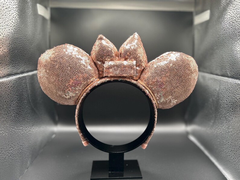May include: Rose gold sequined mouse ear headband with a matching bow. The ears and bow are covered in shimmering sequins. The headband is black and has a black stand.