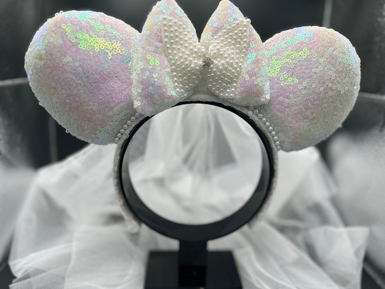Puede incluir: Orejas de Minnie Mouse blancas iridiscentes con un lazo de perlas y un velo de tul blanco. Las orejas est&aacute;n en una diadema negra.
