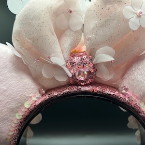 Op de afbeelding: Een haarband met twee grote, zachte, lichtroze pluche oren. Een centrale strik van doorschijnende, glitterende stof is versierd met witte stoffen bloemen en een centrale roze lovertjesknoop met een ronde roze edelsteen. De haarband is bedekt met roze lovertjes.