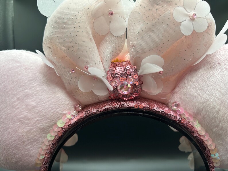 Op de afbeelding: Een haarband met twee grote, zachte, lichtroze pluche oren. Een centrale strik van doorschijnende, glitterende stof is versierd met witte stoffen bloemen en een centrale roze lovertjesknoop met een ronde roze edelsteen. De haarband is bedekt met roze lovertjes.