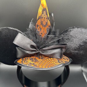 May include: Black mouse ear headband with a witch hat design. The ears are black and plush. The hat is orange with black lace and a black bow. A black feather and tulle accent the hat.