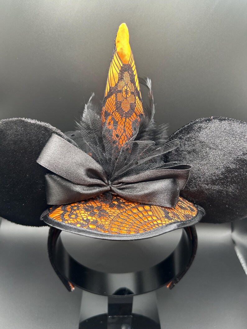 May include: Black mouse ear headband with a witch hat design. The ears are black and plush. The hat is orange with black lace and a black bow. A black feather and tulle accent the hat.