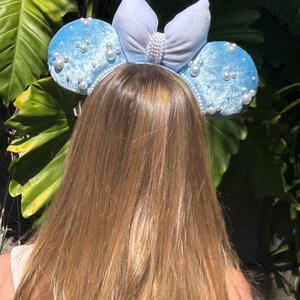 May include: Light blue mouse ear headband with pearl accents and a white bow. The ears are covered in a soft, velvety material. The bow is adorned with a row of pearls. The headband is a fun accessory.
