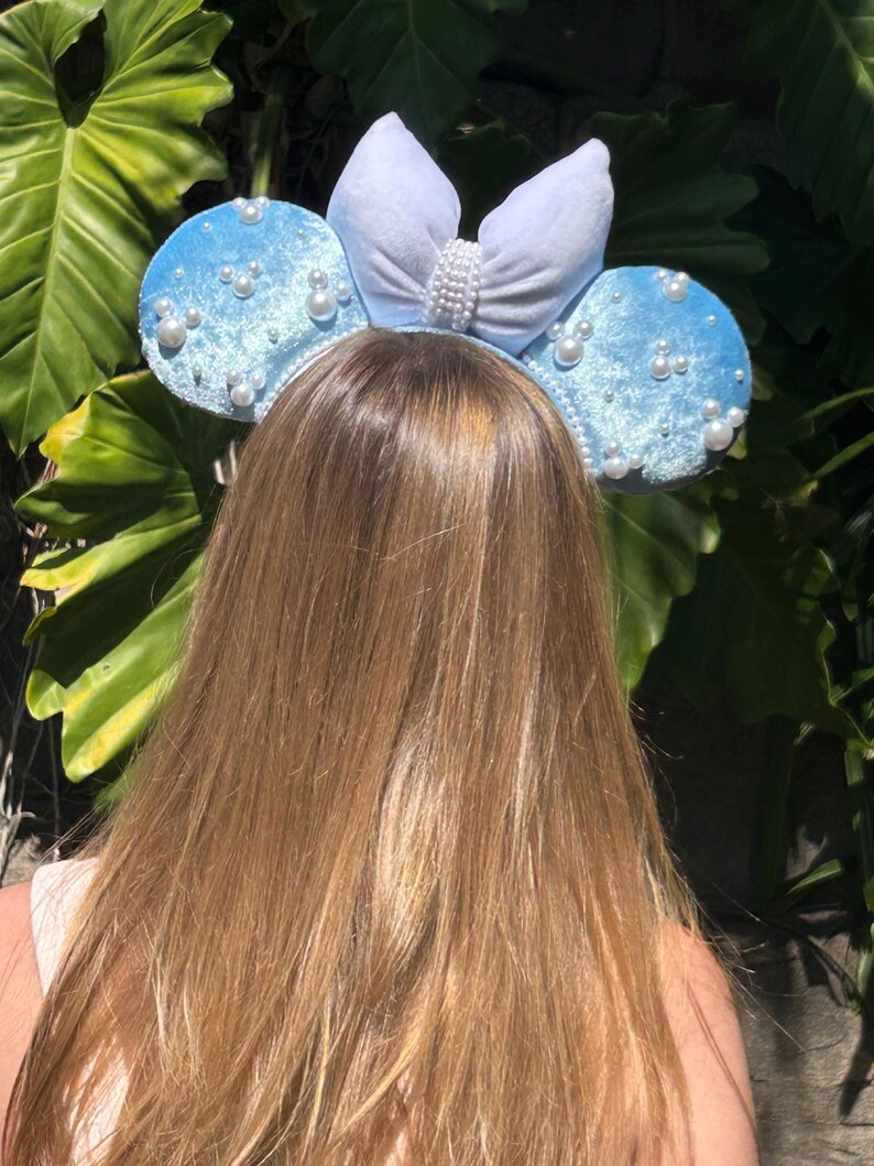 May include: Light blue mouse ear headband with pearl accents and a white bow. The ears are covered in a soft, velvety material. The bow is adorned with a row of pearls. The headband is a fun accessory.