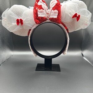 May include: White Minnie Mouse ears with a red sequined bow and white lace accents. The bow has a white pearl center.