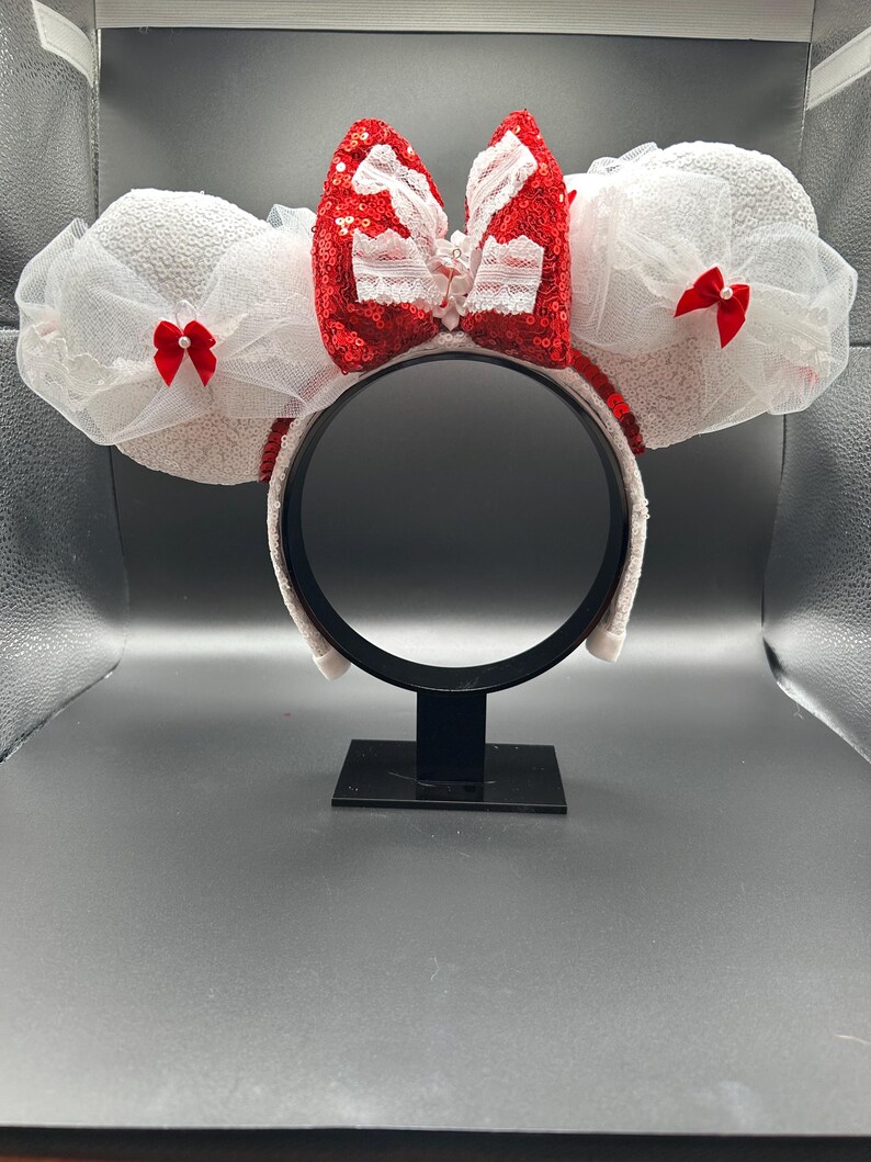 May include: White Minnie Mouse ears with a red sequined bow and white lace accents. The bow has a white pearl center.
