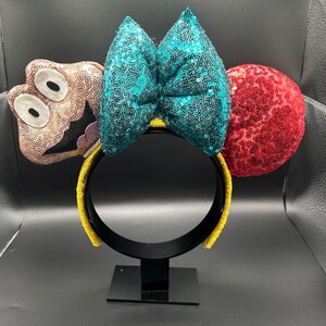 May include: A whimsical headband featuring a teal sequined bow, a red sequined ear, and a character-shaped ear with a smiling face. The headband has a yellow band and is displayed on a black stand.
