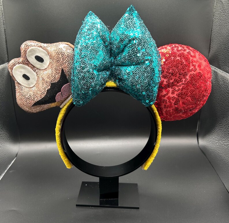 May include: A whimsical headband featuring a teal sequined bow, a red sequined ear, and a character-shaped ear with a smiling face. The headband has a yellow band and is displayed on a black stand.
