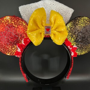 May include: A headband featuring two circular ears covered in red and gold sequins, accented with small red fabric roses at the base. A large yellow sequined bow sits in the center, adorned with a small rectangular charm featuring a red heart.