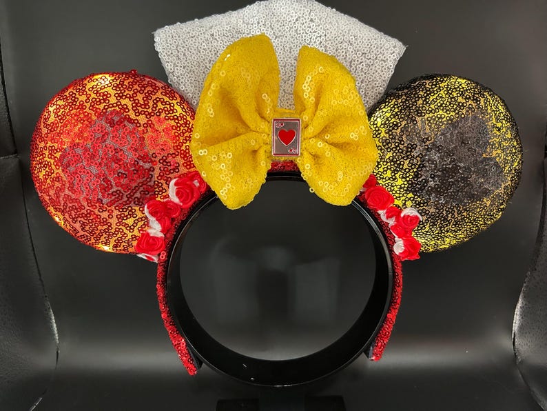 May include: A headband featuring two circular ears covered in red and gold sequins, accented with small red fabric roses at the base. A large yellow sequined bow sits in the center, adorned with a small rectangular charm featuring a red heart.
