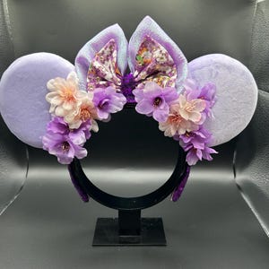 Puede incluir: Una diadema con dos orejas grandes y redondas de felpa color morado claro. Un lazo central muestra un patr&oacute;n floral con detalles dorados. Racimos de flores artificiales en tonos rosa claro, lavanda y morado oscuro decoran la parte frontal.