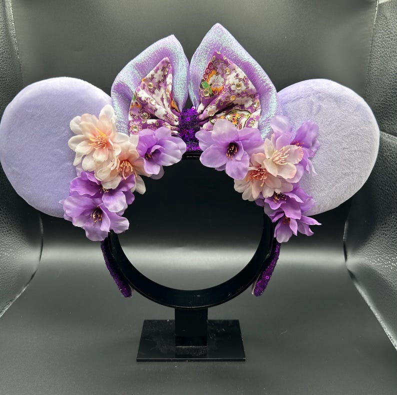 Puede incluir: Una diadema con dos orejas grandes y redondas de felpa color morado claro. Un lazo central muestra un patr&oacute;n floral con detalles dorados. Racimos de flores artificiales en tonos rosa claro, lavanda y morado oscuro decoran la parte frontal.