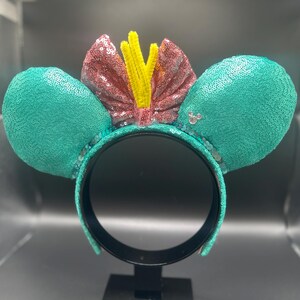 May include: A teal sequined headband with large mouse ears. The ears are covered in teal sequins. A pink sequined bow with yellow accents sits between the ears. The headband is black.
