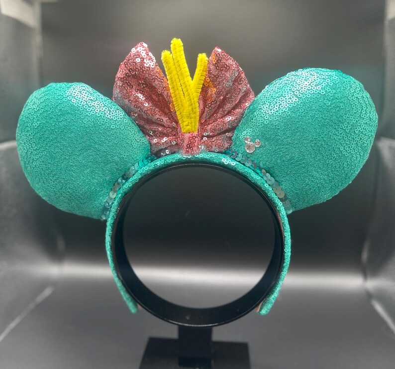 May include: A teal sequined headband with large mouse ears. The ears are covered in teal sequins. A pink sequined bow with yellow accents sits between the ears. The headband is black.