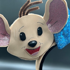 May include: A brown mouse character headband with pink ears, a black nose, and a red mouth. The headband has a blue sequined bow.