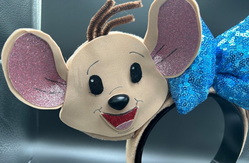 May include: A brown mouse character headband with pink ears, a black nose, and a red mouth. The headband has a blue sequined bow.