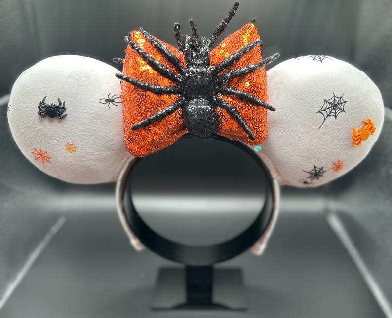 May include: Halloween-themed mouse ears. The white plush ears are decorated with spiders, spiderwebs, and orange accents. A large orange sequined bow with a black glitter spider is centered on the black headband.