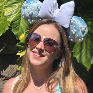 May include: A light blue headband with two round ears covered in small white pearls. A large, light blue bow is centered between the ears. The headband is worn by a person wearing sunglasses and a light blue top.