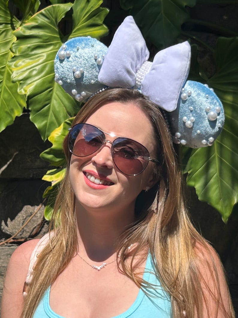 May include: A light blue headband with two round ears covered in small white pearls. A large, light blue bow is centered between the ears. The headband is worn by a person wearing sunglasses and a light blue top.