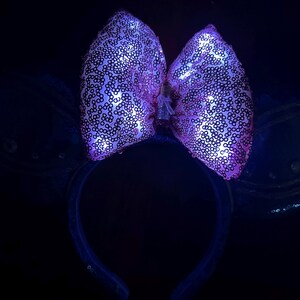 May include: A light-up headband with a large, sequined bow in shades of pink and purple. The bow is illuminated with small lights, creating a glowing effect. The headband and attached wings are dark blue, with a few sequins.