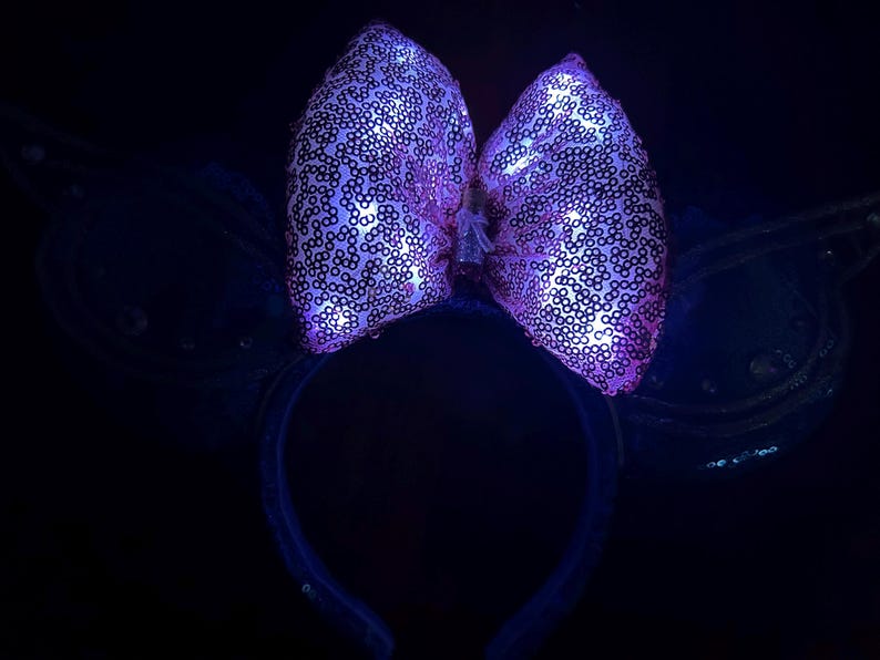 May include: A light-up headband with a large, sequined bow in shades of pink and purple. The bow is illuminated with small lights, creating a glowing effect. The headband and attached wings are dark blue, with a few sequins.