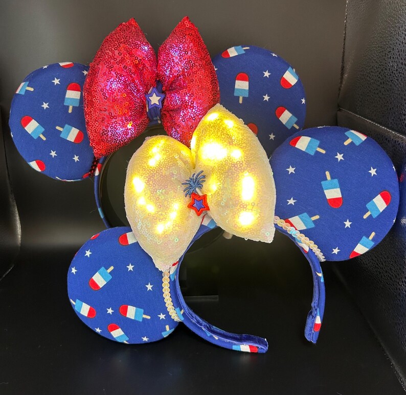 K&ouml;nnte beinhalten: Ein festliches Stirnband mit Mauseohren, mit einer roten Pailletten-Schleife und einer wei&szlig;en Pailletten-Schleife mit Lichtern. Die Ohren sind blau mit einem Eis- und Sternenmuster. Das Stirnband ist blau.