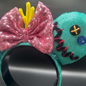 May include: Teal sequined mouse ears with a pink sequined bow and yellow pipe cleaner accents. One ear features black stitching and a button. The headband is black.