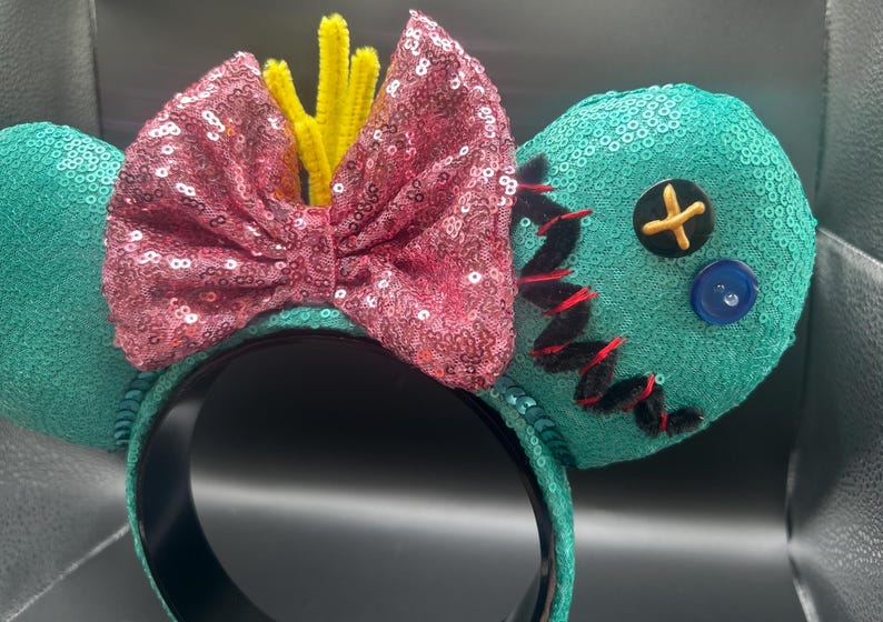 May include: Teal sequined mouse ears with a pink sequined bow and yellow pipe cleaner accents. One ear features black stitching and a button. The headband is black.