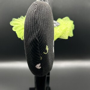 May include: Black headband with a ribbed texture, adorned with a lime green tulle accent. The headband features a decorative element with a black and silver design. The design includes a small, embroidered detail in white and lime green.