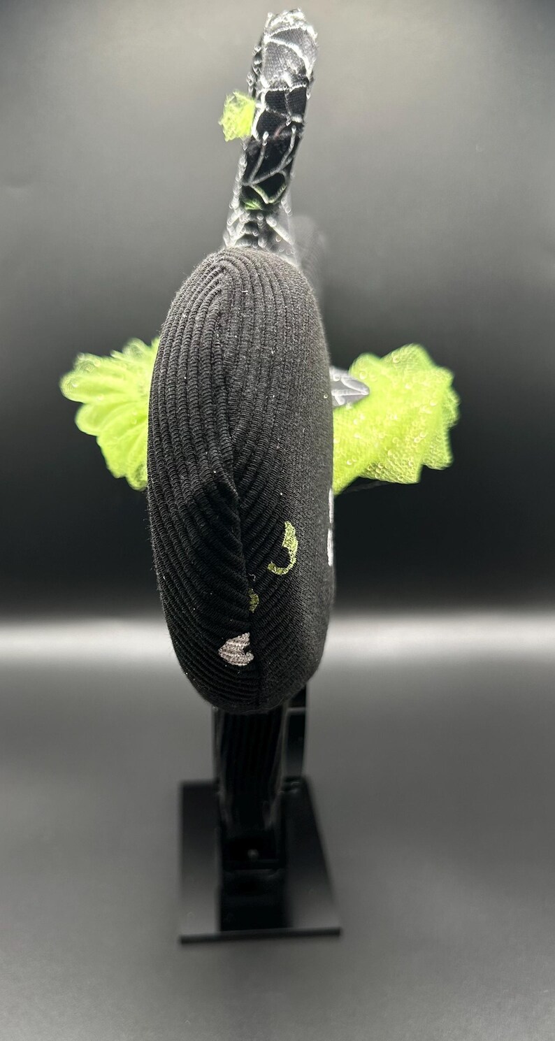 May include: Black headband with a ribbed texture, adorned with a lime green tulle accent. The headband features a decorative element with a black and silver design. The design includes a small, embroidered detail in white and lime green.