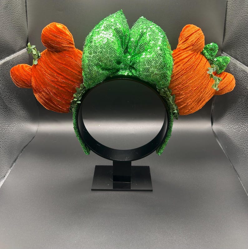 May include: A festive headband featuring a large green sequined bow and two orange, textured, Mickey Mouse-shaped accents. The headband is black, with green accents, and is displayed on a black stand.
