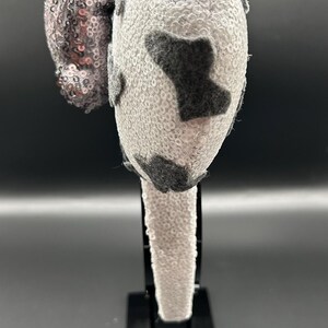 May include: A headband featuring two round ears covered in white sequins with dark grey felt patches. A large, upright bow covered in shimmering silver sequins is attached between the ears. The headband itself is also covered in white sequins.