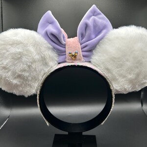 May include: A headband with white fluffy ears, a lavender bow, and a pink center adorned with a small gold embellishment. The headband is white with a silver trim. The ears are round and plush.