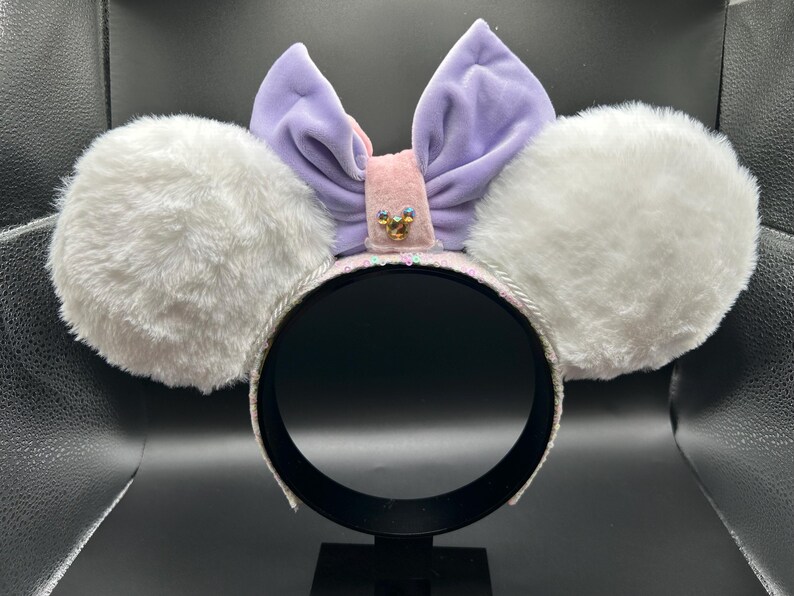 May include: A headband with white fluffy ears, a lavender bow, and a pink center adorned with a small gold embellishment. The headband is white with a silver trim. The ears are round and plush.