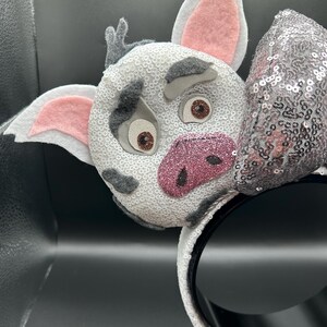 May include: A headband featuring a character face covered in white sequins with grey felt patches, pink felt inner ears, and a glittery pink snout. The character has brown eyes and grey felt eyebrows. A silver sequined bow sits to the right side.